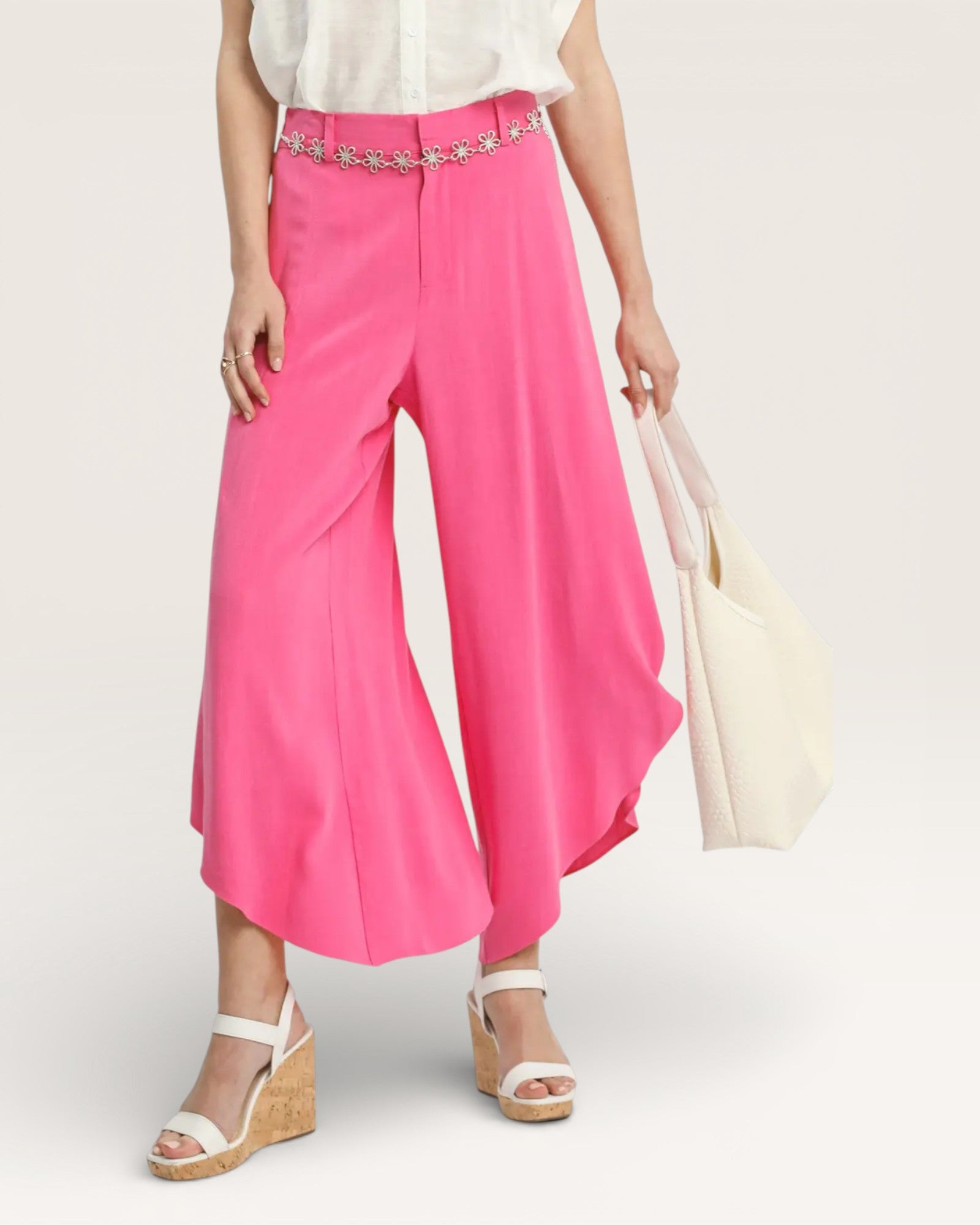 Person wearing bright pink wide-leg pants with a white top and sandals, holding a beige bag on a light gray background.