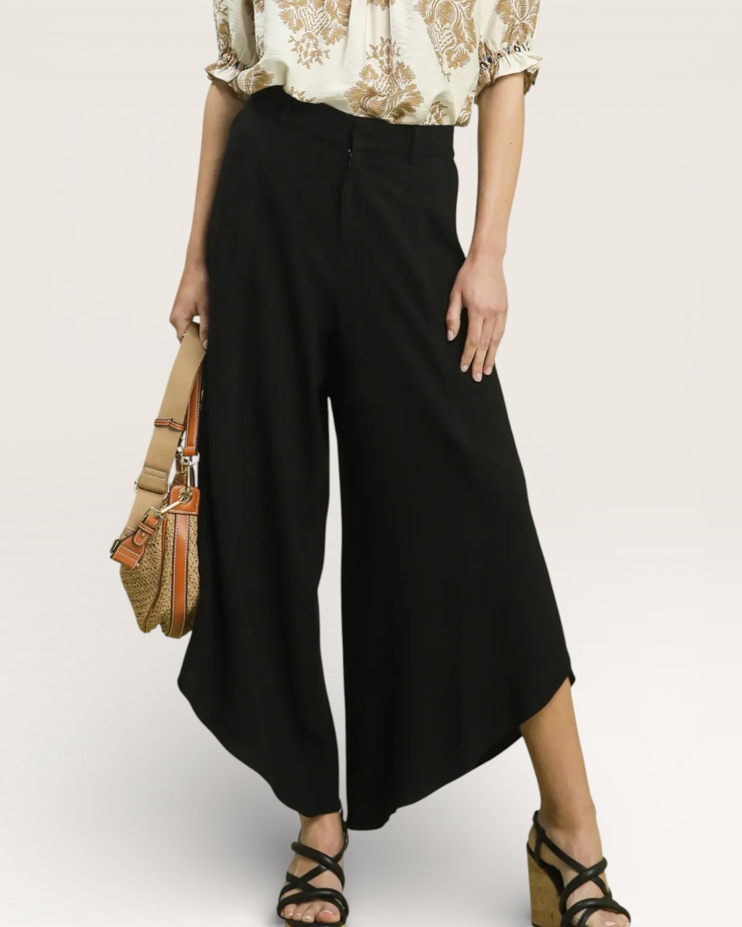 Person wearing black wide-leg pants with a beige top and holding a straw bag on a plain background