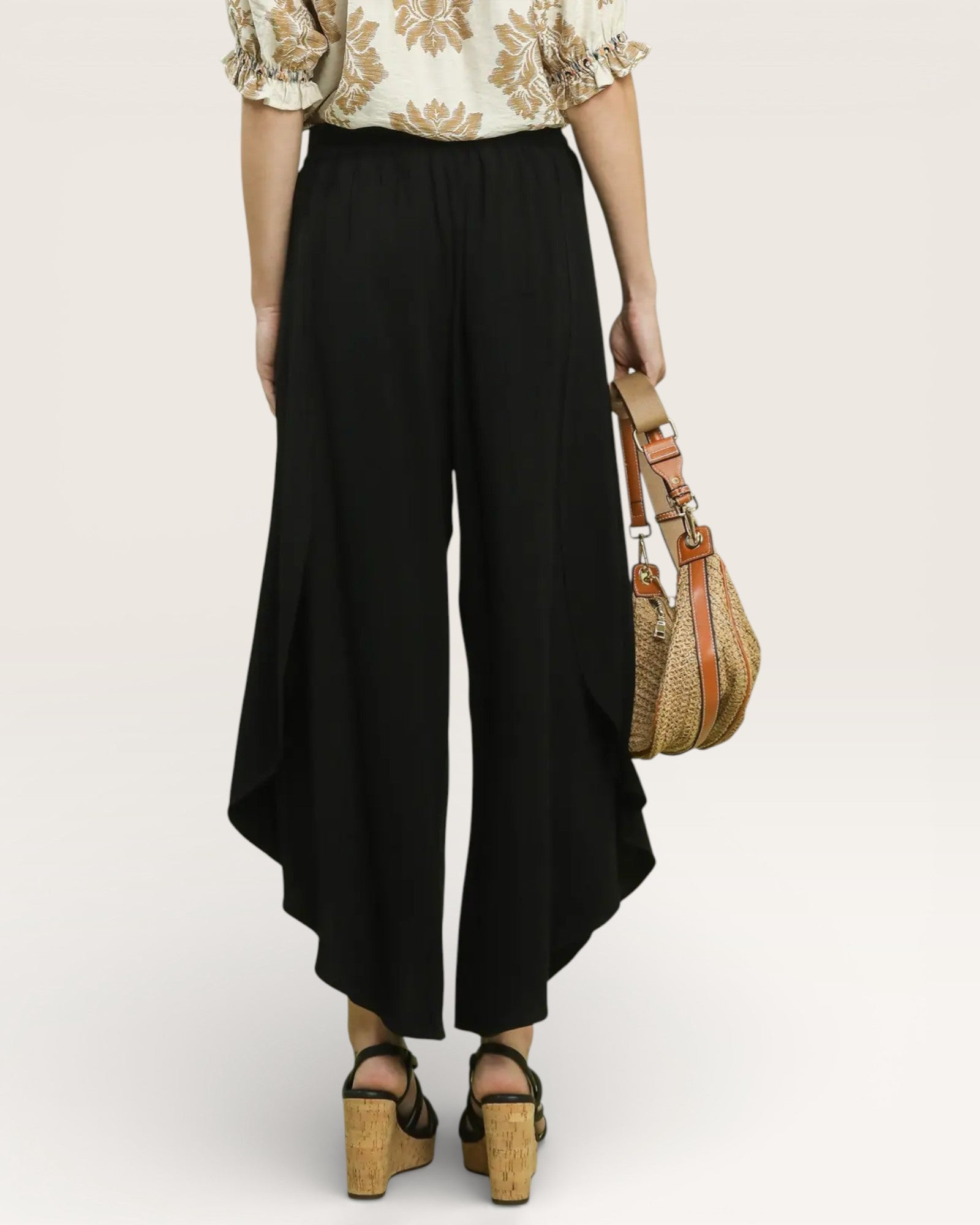 Person wearing black wide-leg pants with a patterned top and holding a straw bag on a white background
