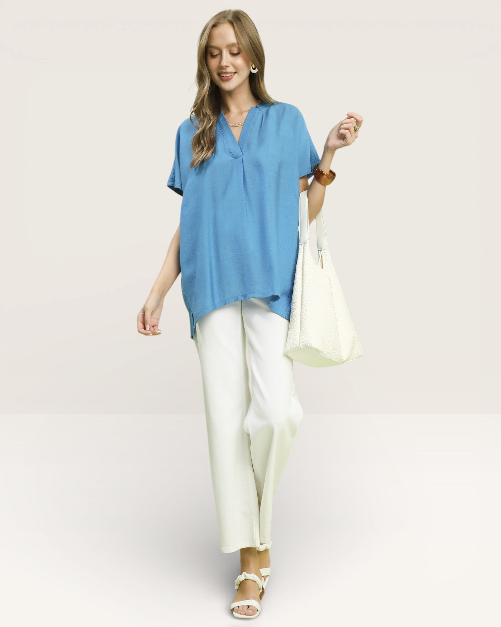 Woman wearing a blue top and white pants holding a white bag on a light background