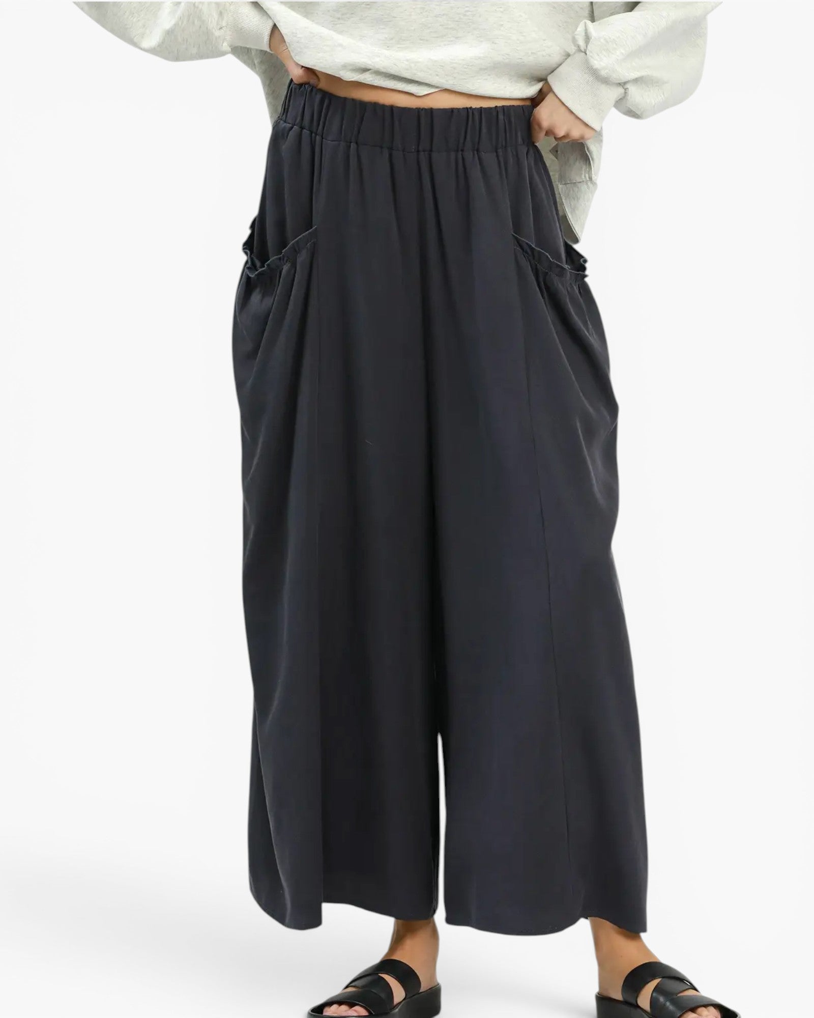 A person wearing loose, wide-leg cropped pants in a very dark navy or near-black color, paired with a light gray top and black slide sandals, against a plain light background.