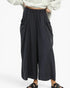 A person wearing loose, wide-leg cropped pants in a very dark navy or near-black color, paired with a light gray top and black slide sandals, against a plain light background.