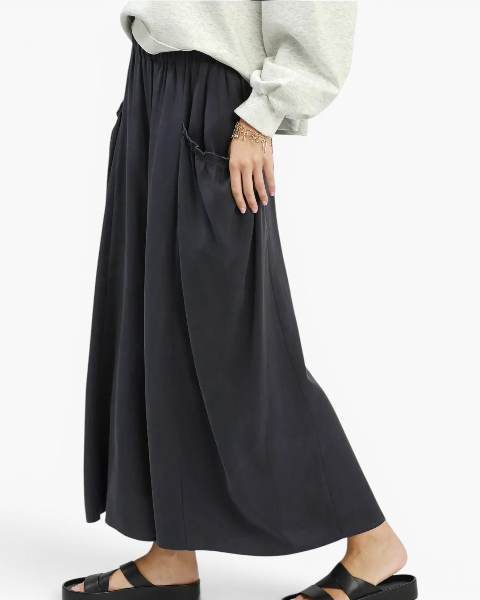A person wearing a loose, flowing black maxi skirt with side pockets, paired with a light gray sweatshirt and black slide sandals, shown against a plain light gray background.