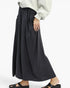 A person wearing a loose, flowing black maxi skirt with side pockets, paired with a light gray sweatshirt and black slide sandals, shown against a plain light gray background.