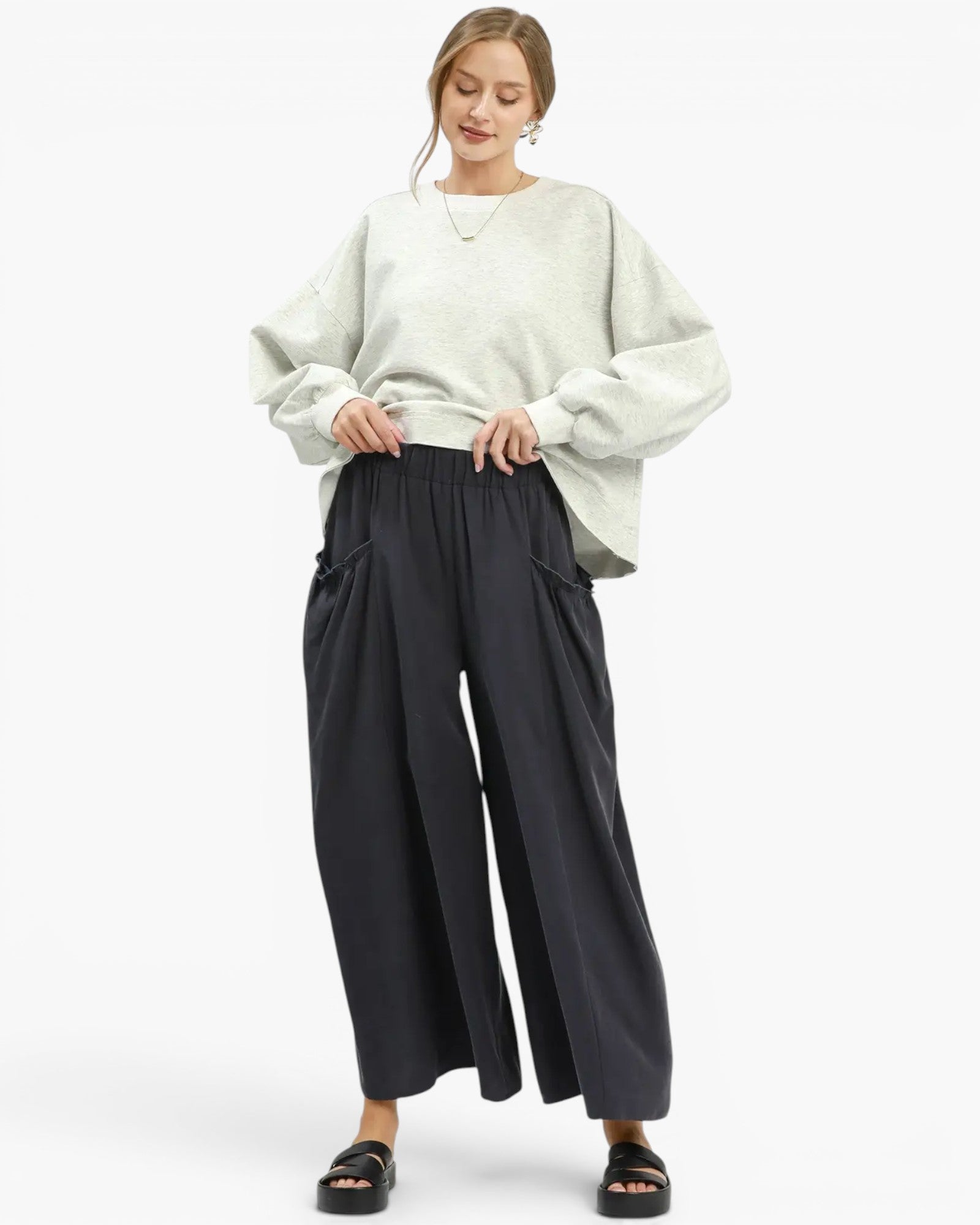 A woman modeling a light sage green oversized sweatshirt with voluminous sleeves and black wide-leg cropped pants, paired with black slide sandals, against a plain white studio background.
