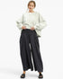 A woman modeling a light sage green oversized sweatshirt with voluminous sleeves and black wide-leg cropped pants, paired with black slide sandals, against a plain white studio background.