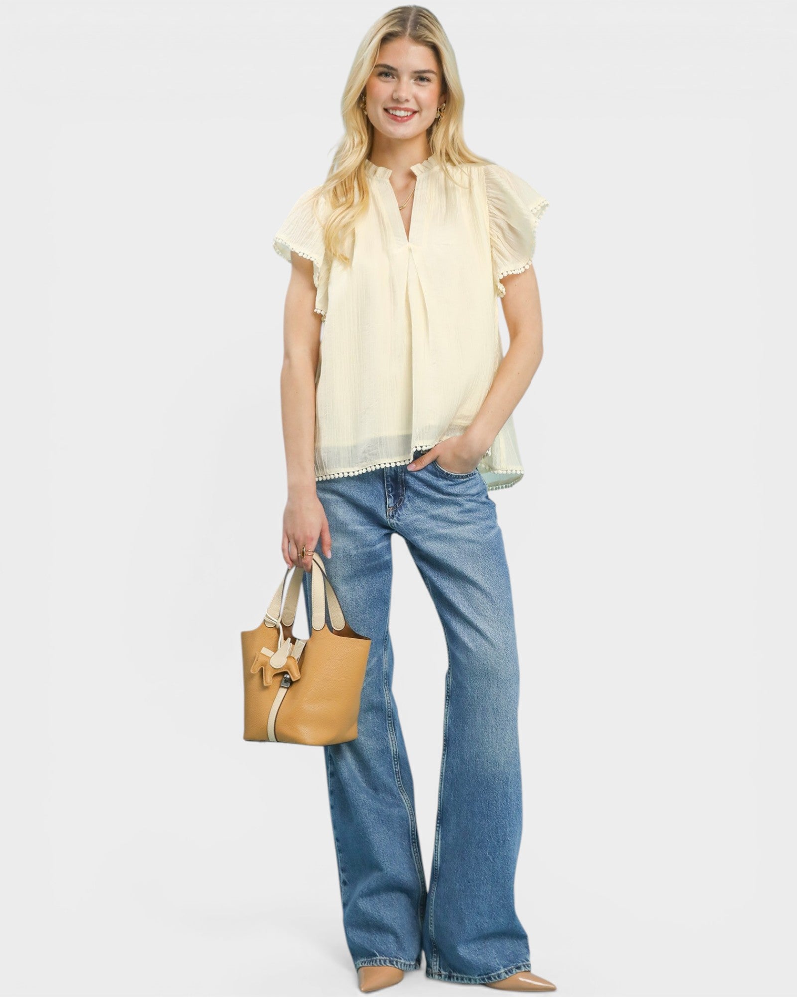 Woman wearing a light yellow blouse and blue jeans, holding a tan handbag on a white background