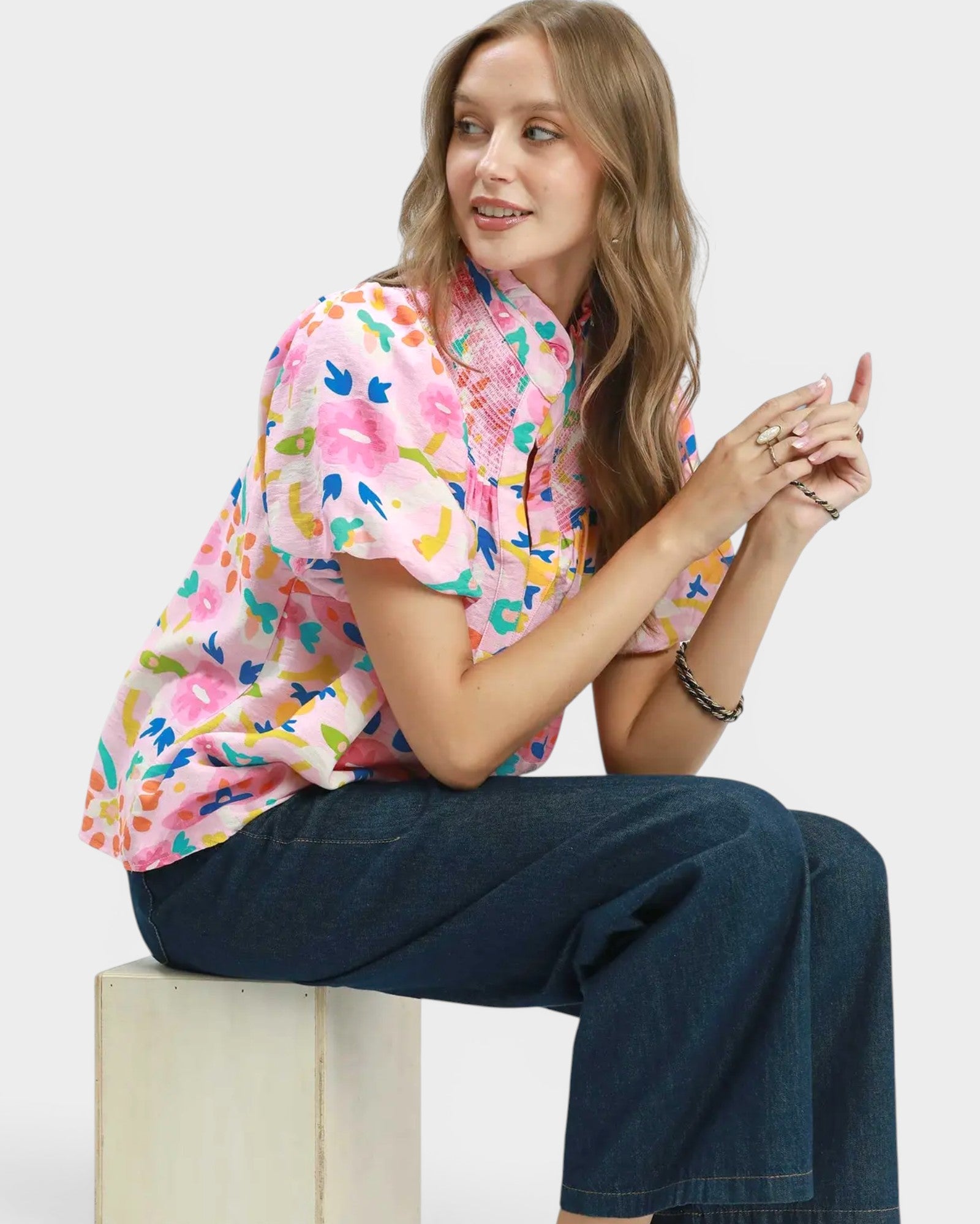 Woman wearing a colorful floral blouse and blue jeans sitting on a white block.