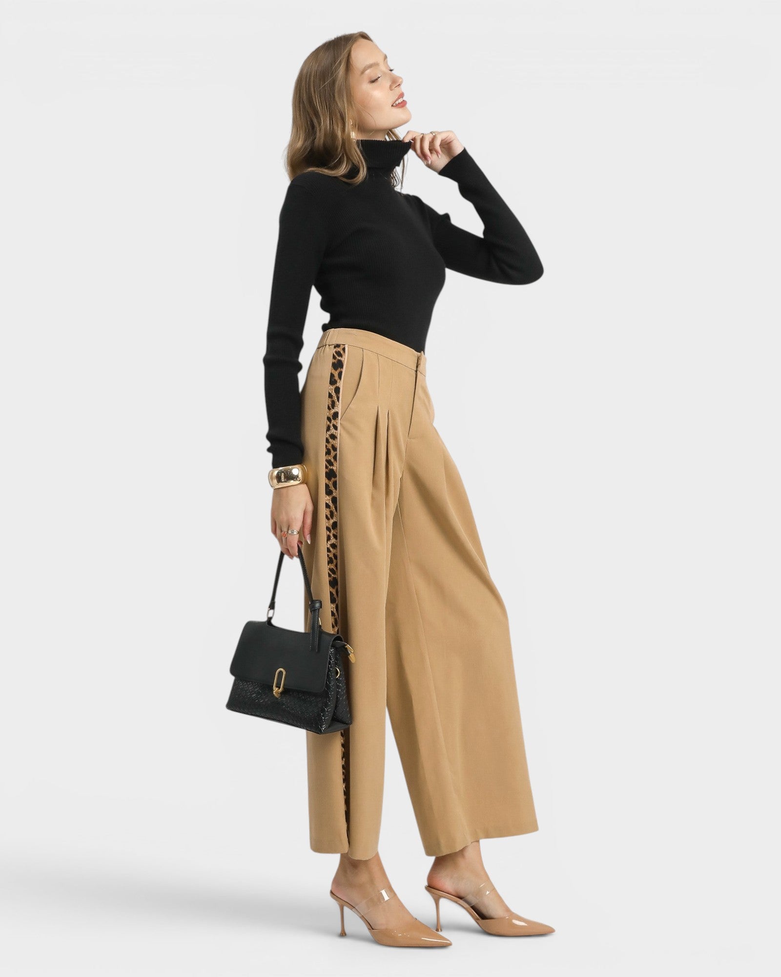 Woman wearing a black top and beige pants with lace details, holding a black handbag on a white background.