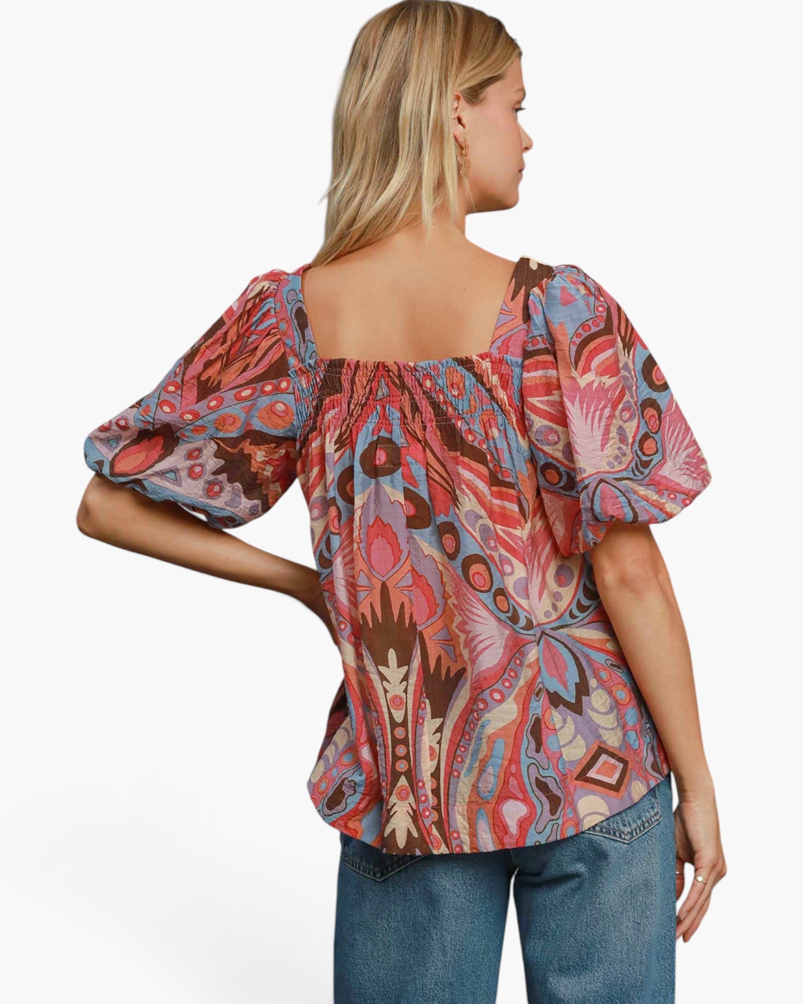 A woman shown from the back wearing a colorful puff-sleeve top with an abstract paisley-style print in pink, blue, brown, and beige, paired with blue jeans against a plain white studio background.