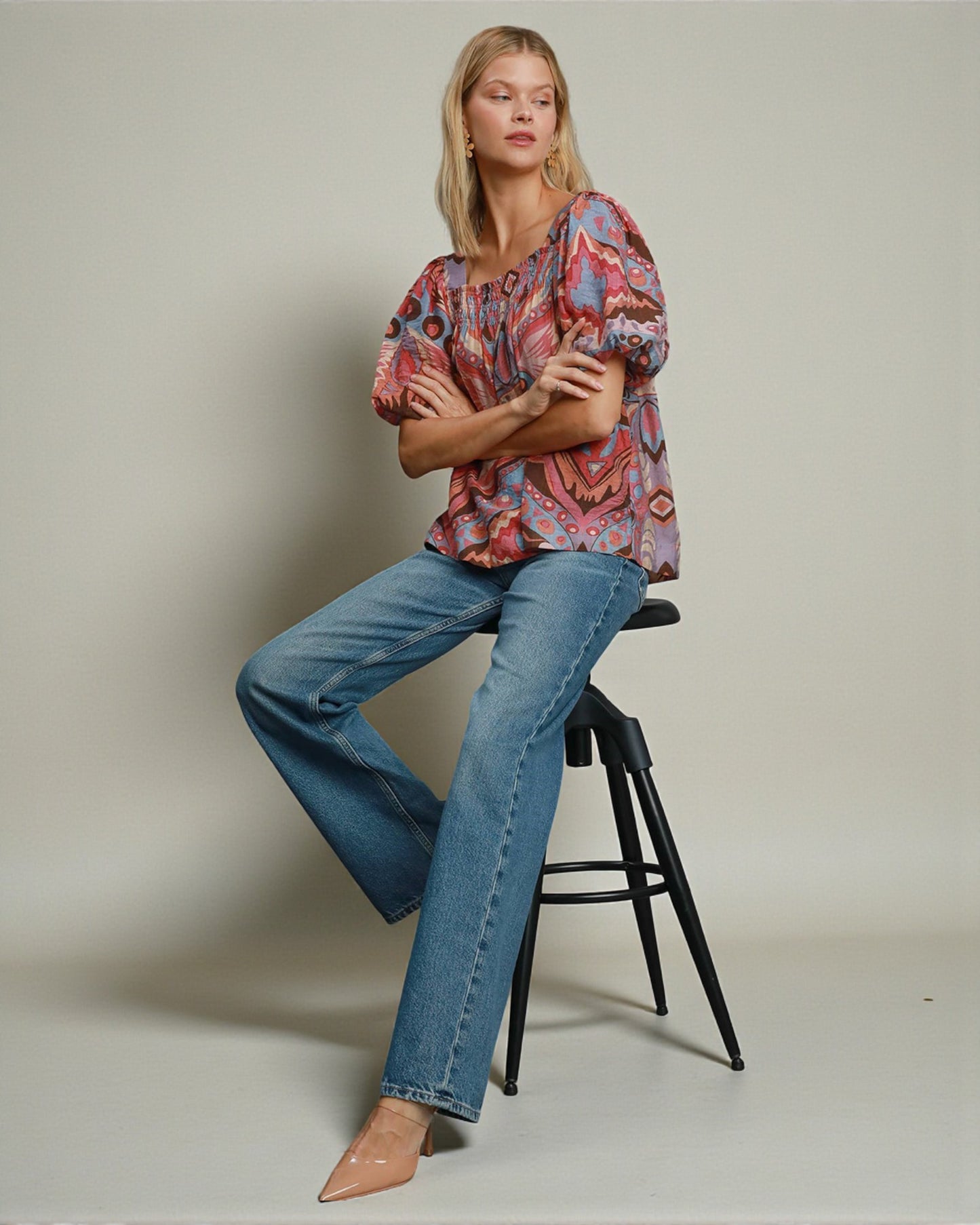 Woman sitting on a stool wearing a patterned top and blue jeans against a plain background