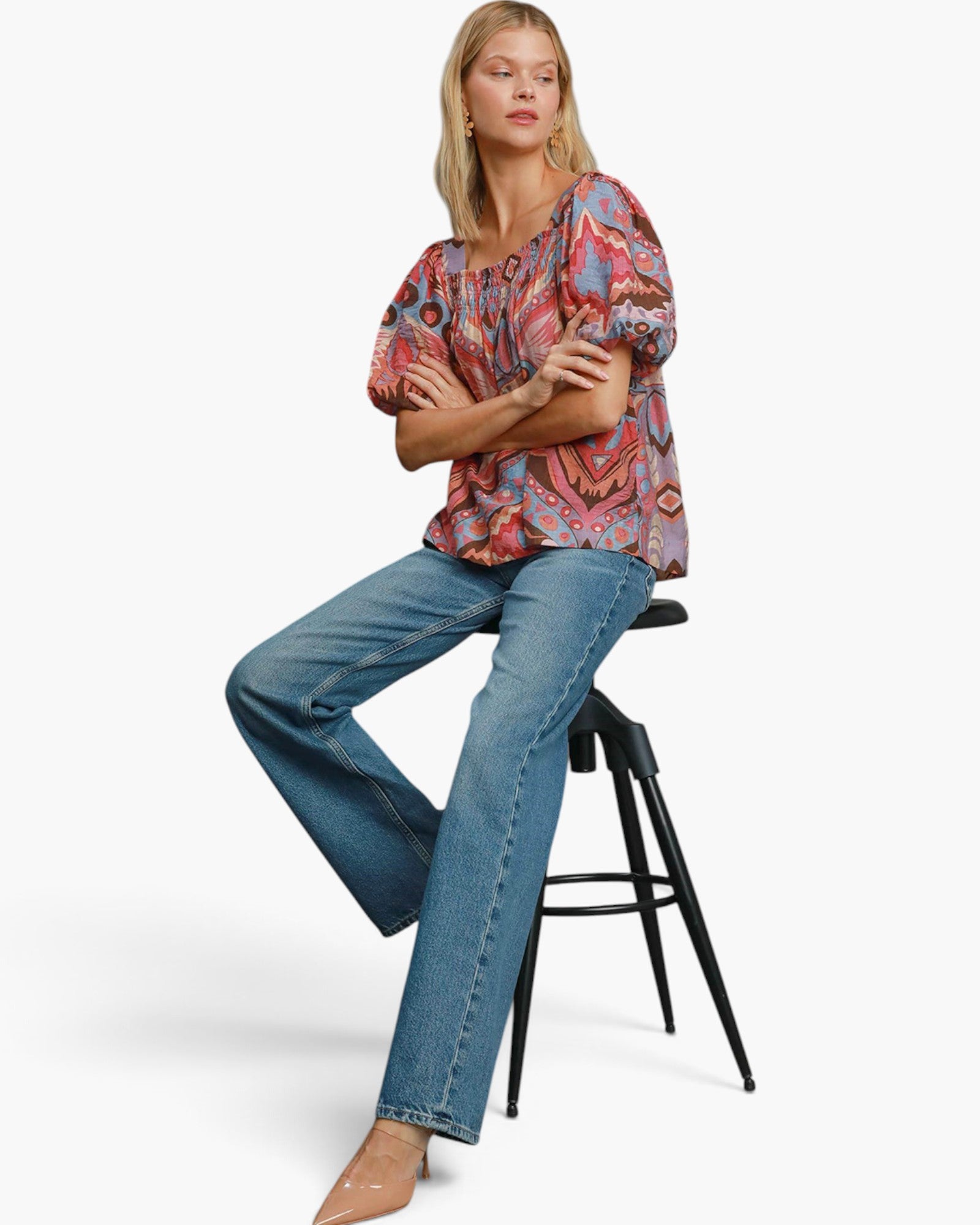 A woman sits on a black stool wearing a colorful patterned blouse and blue jeans against a plain light gray studio background.
