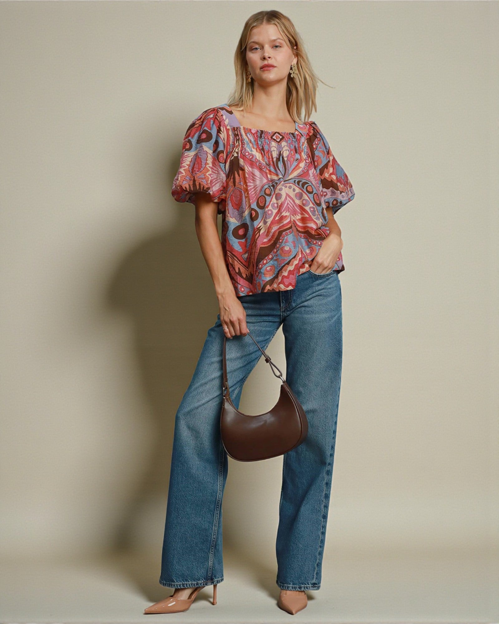 Woman wearing a patterned top and jeans, holding a brown handbag against a beige background