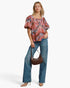 A model wearing a colorful, patterned puff-sleeve blouse with blue wide-leg jeans, holding a brown crescent handbag, and wearing beige heels against a plain white background.