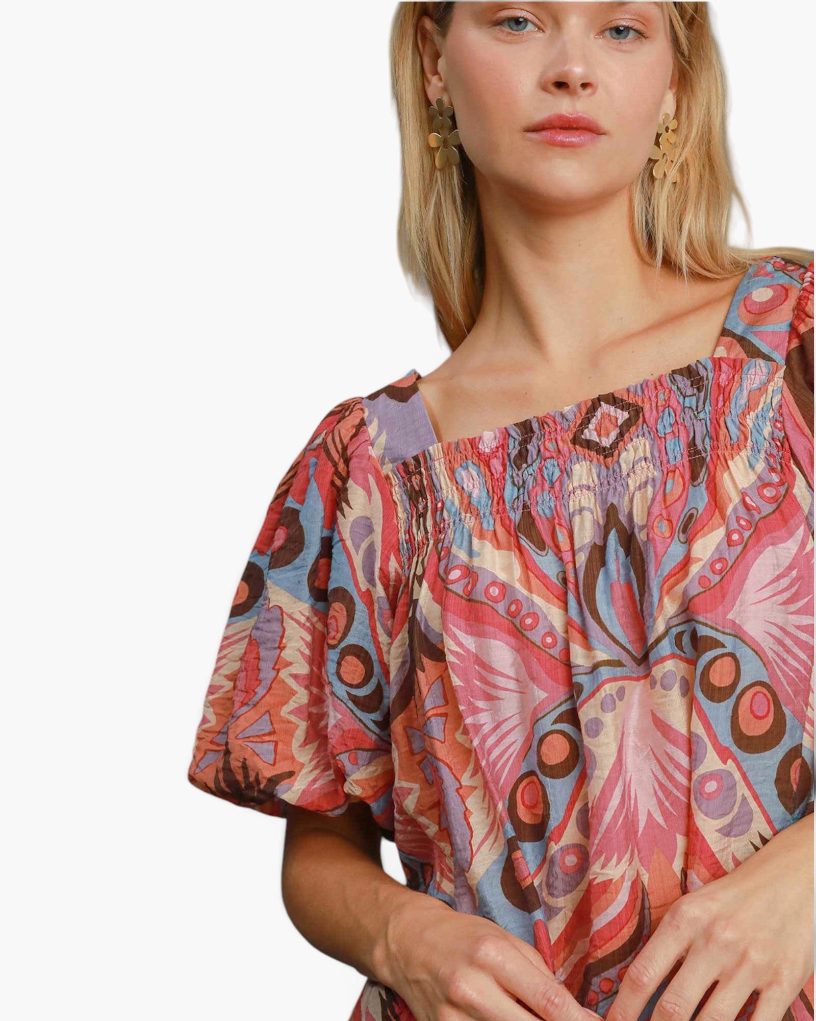 A close-up of a woman wearing a colorful, patterned short-sleeve top with a smocked neckline. The top features a busy abstract print in pink, orange, blue, and brown tones.