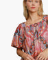 A close-up of a woman wearing a colorful, patterned short-sleeve top with a smocked neckline. The top features a busy abstract print in pink, orange, blue, and brown tones.