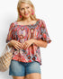 A woman modeling a bohemian-style short-sleeve top with a colorful paisley and floral pattern in red, blue, purple, and brown, paired with denim shorts and a woven straw bag against a plain white background.