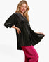 A woman modeling a black long-sleeve tunic top with a small white polka dot pattern, paired with bright pink pants, against a plain white studio background.