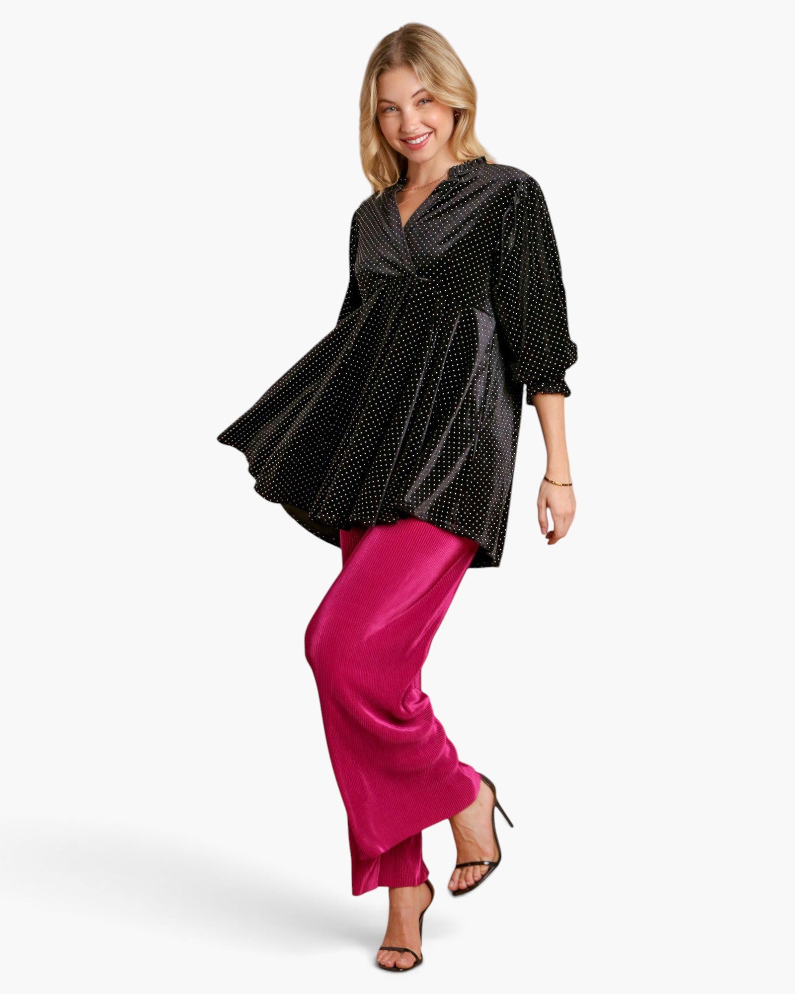 A woman modeling a black long-sleeve top with a small white polka dot pattern and bright magenta satin pants against a light gray studio background.