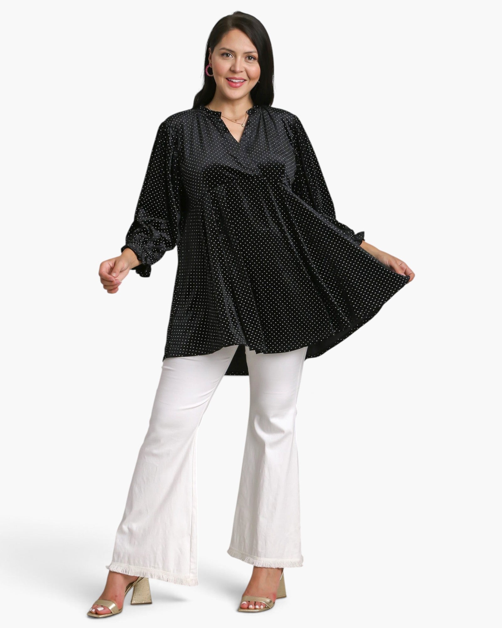 A woman modeling a black long-sleeve top with a small white polka dot pattern, paired with white flared pants and beige heeled sandals, against a plain white studio background.