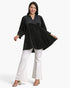 A woman modeling a black long-sleeve top with a small white polka dot pattern, paired with white flared pants and beige heeled sandals, against a plain white studio background.