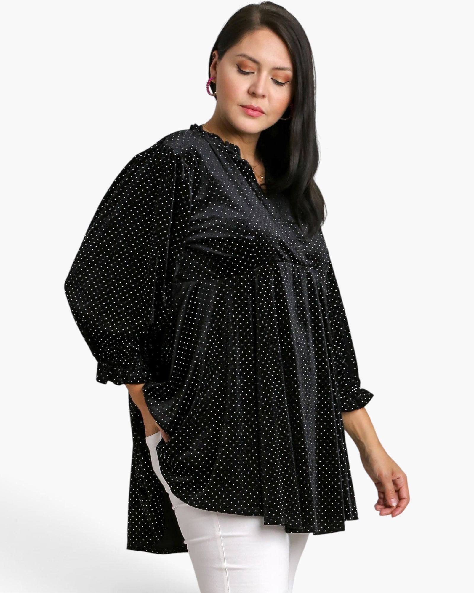 A woman wearing a black long-sleeve tunic top with a white polka dot pattern, paired with white pants, photographed against a clean white studio background.