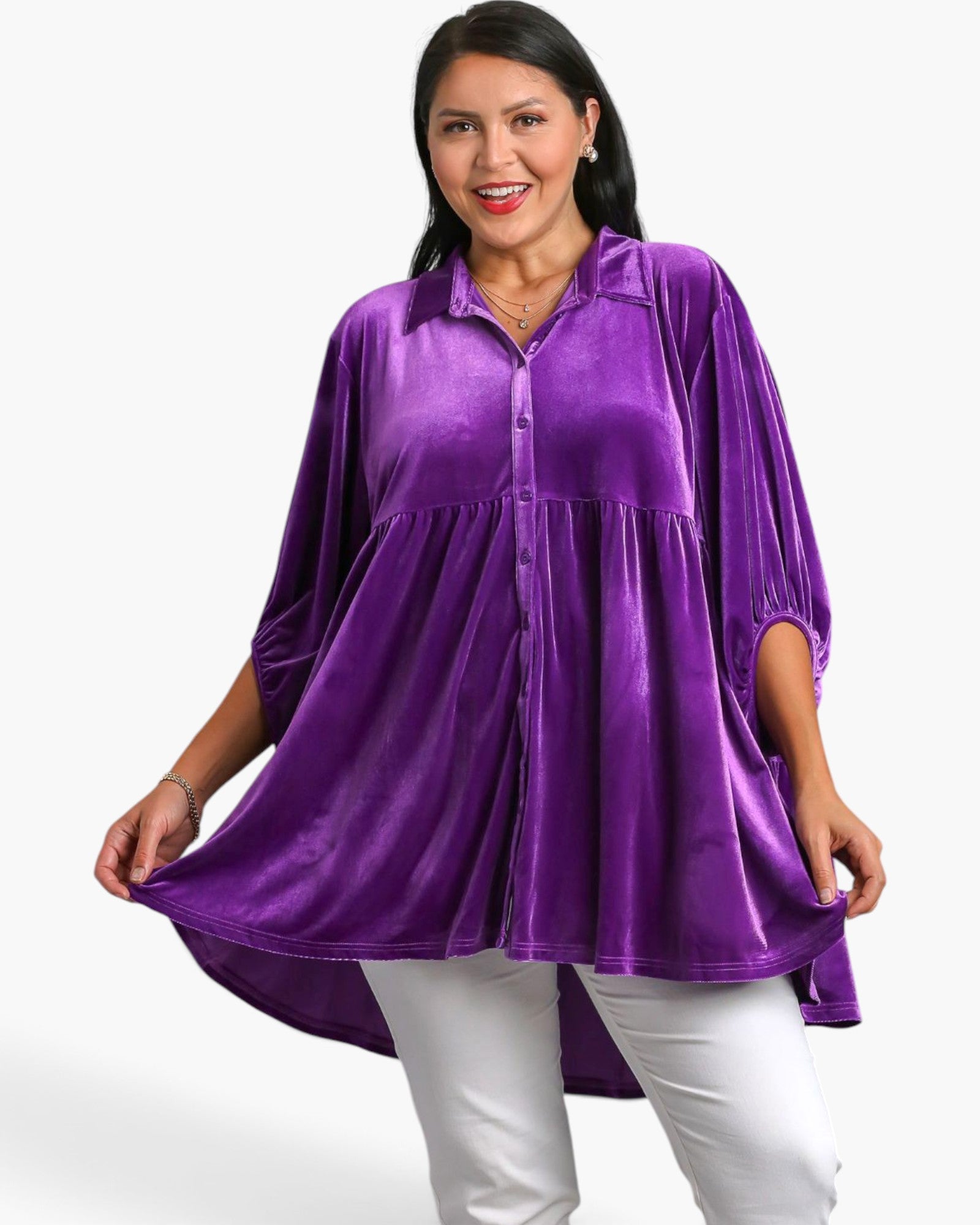 A woman modeling a purple velvet long-sleeve button-up top with a tiered peplum waist, paired with white pants, against a plain white background.