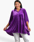 A woman modeling a purple velvet long-sleeve button-up top with a tiered peplum waist, paired with white pants, against a plain white background.