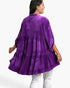 A woman shown from the back wearing a purple tiered velvet tunic top with white pants, photographed against a plain white studio background.