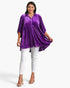 A woman modeling a purple velvet long-sleeve button-up tunic top with white pants and metallic heeled sandals, set against a plain light gray studio background.