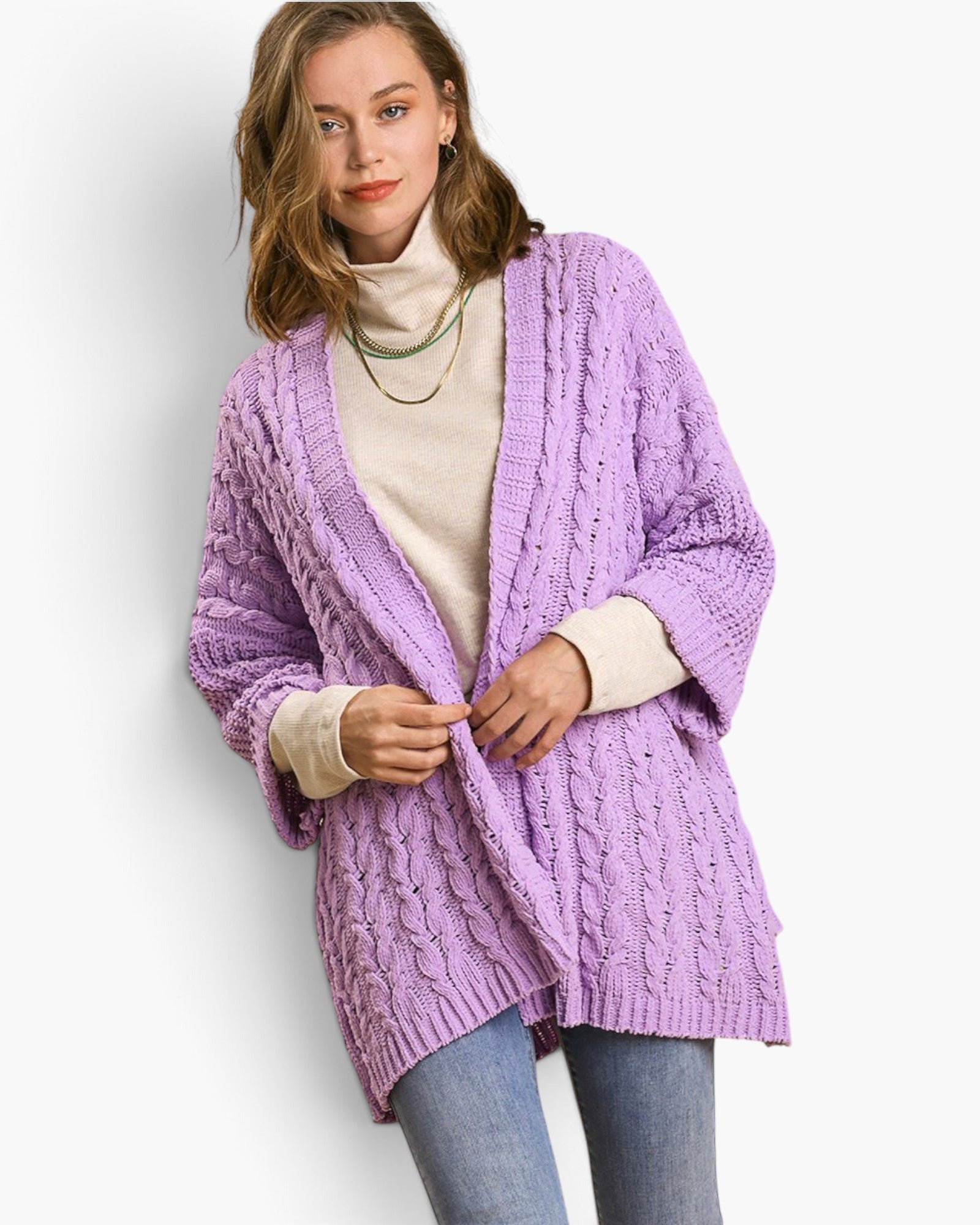 A woman wearing a chunky cable-knit cardigan in light purple over a cream turtleneck sweater and blue jeans, standing against a plain white studio background.