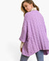 A woman wearing a chunky cable-knit lavender cardigan over a cream turtleneck and blue jeans, photographed from the back against a plain white studio background.