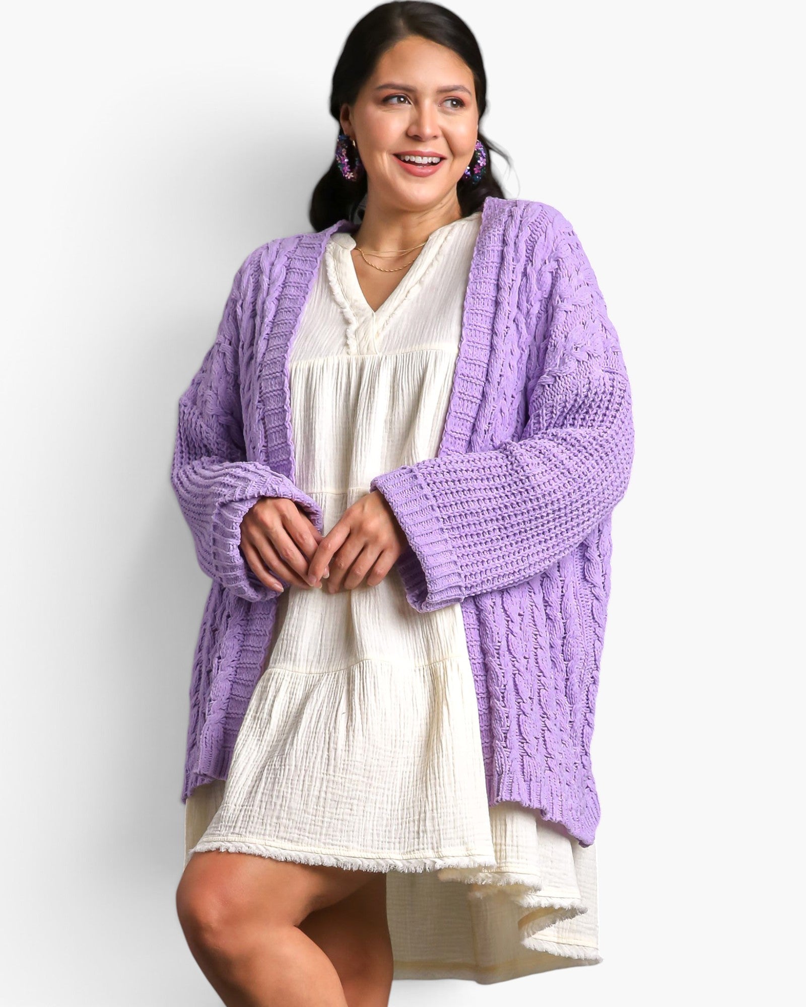 A woman modeling a lavender purple chunky cable-knit cardigan over a cream-colored tiered dress, set against a plain light gray studio background.