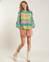 Person wearing a colorful striped sweater with pom-poms, pink shorts, and white boots on a light background