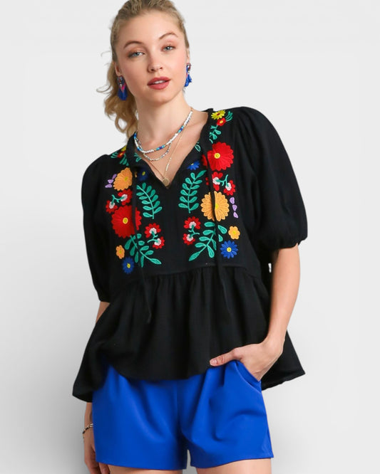 Woman wearing a black top with colorful floral embroidery and blue shorts on a white background