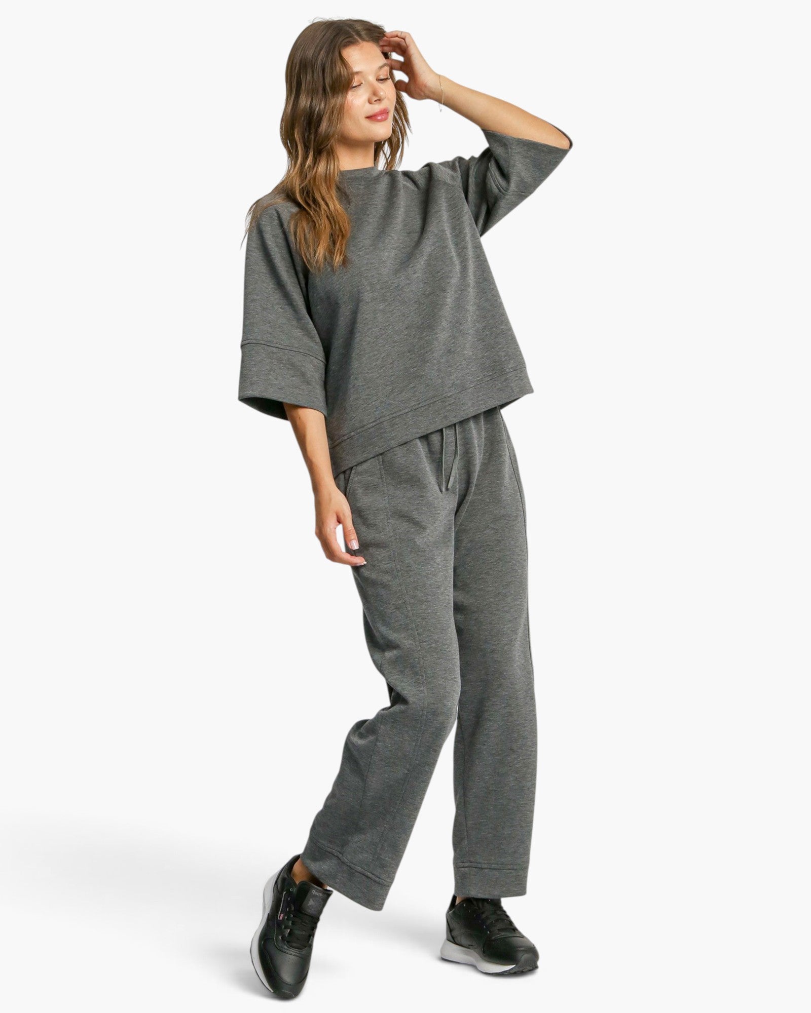 A woman modeling a dark gray heathered lounge set consisting of a cropped short-sleeve top and matching drawstring sweatpants, paired with black sneakers, against a light gray studio background.