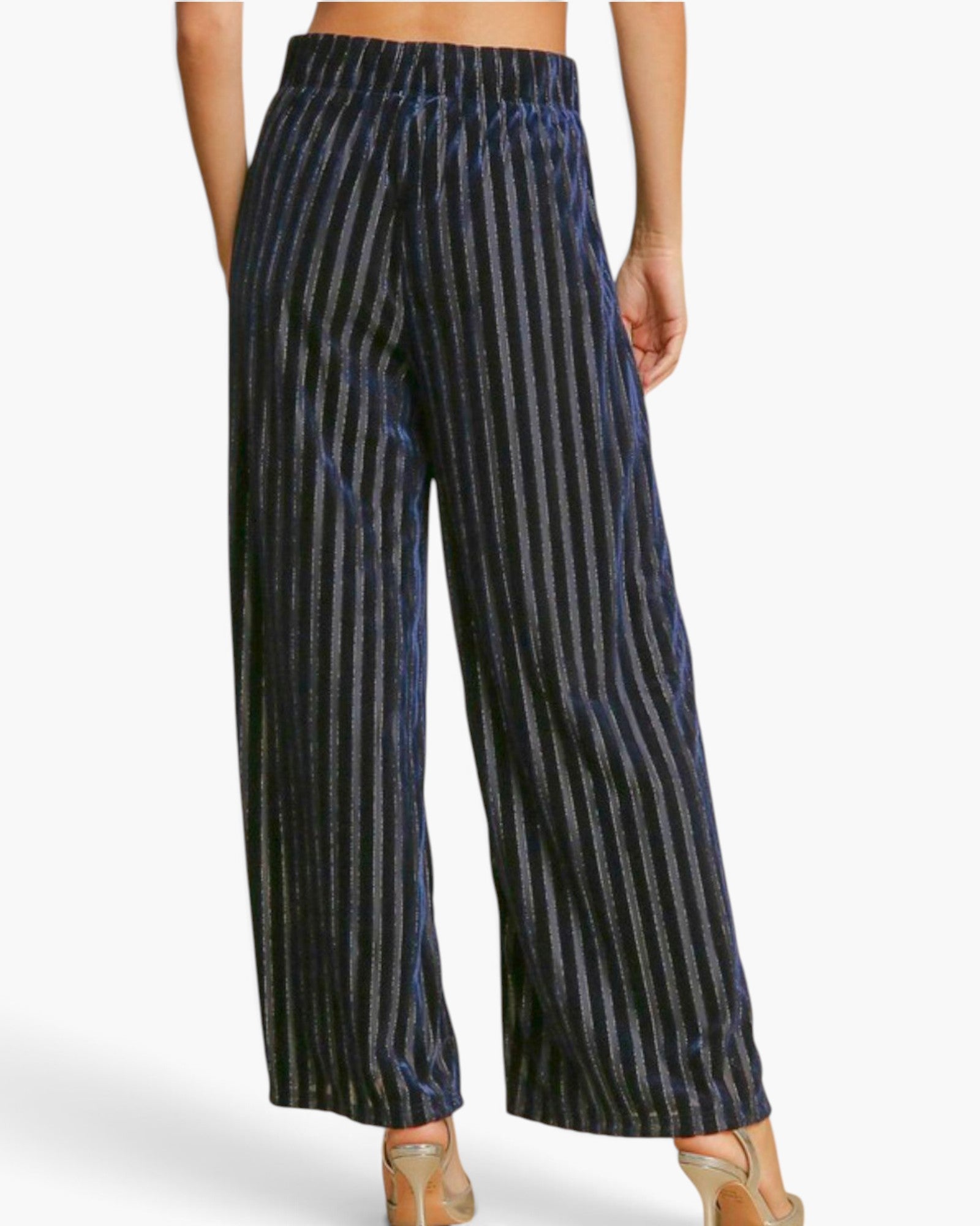 A model from the back wearing high-waisted, wide-leg trousers with a vertical metallic stripe pattern in navy blue and silver, paired with clear high-heeled sandals.
