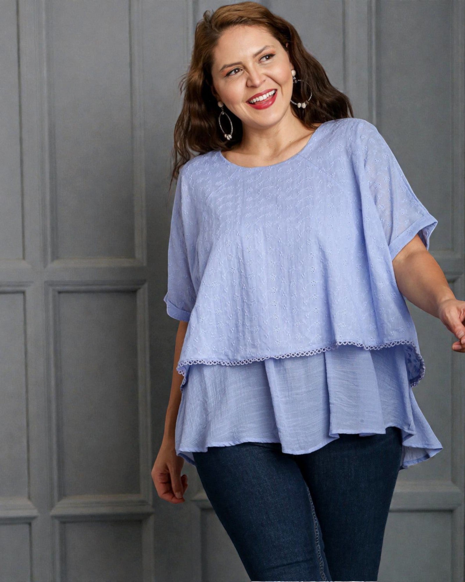 Woman wearing a light blue layered top and dark jeans against a gray background