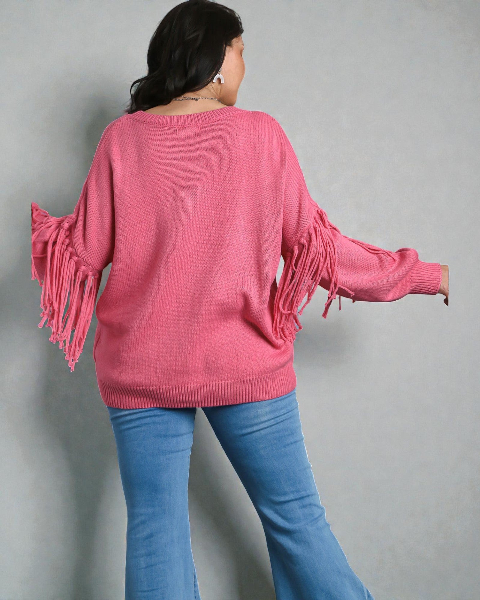 Person wearing a pink sweater with fringe details and blue jeans against a gray background