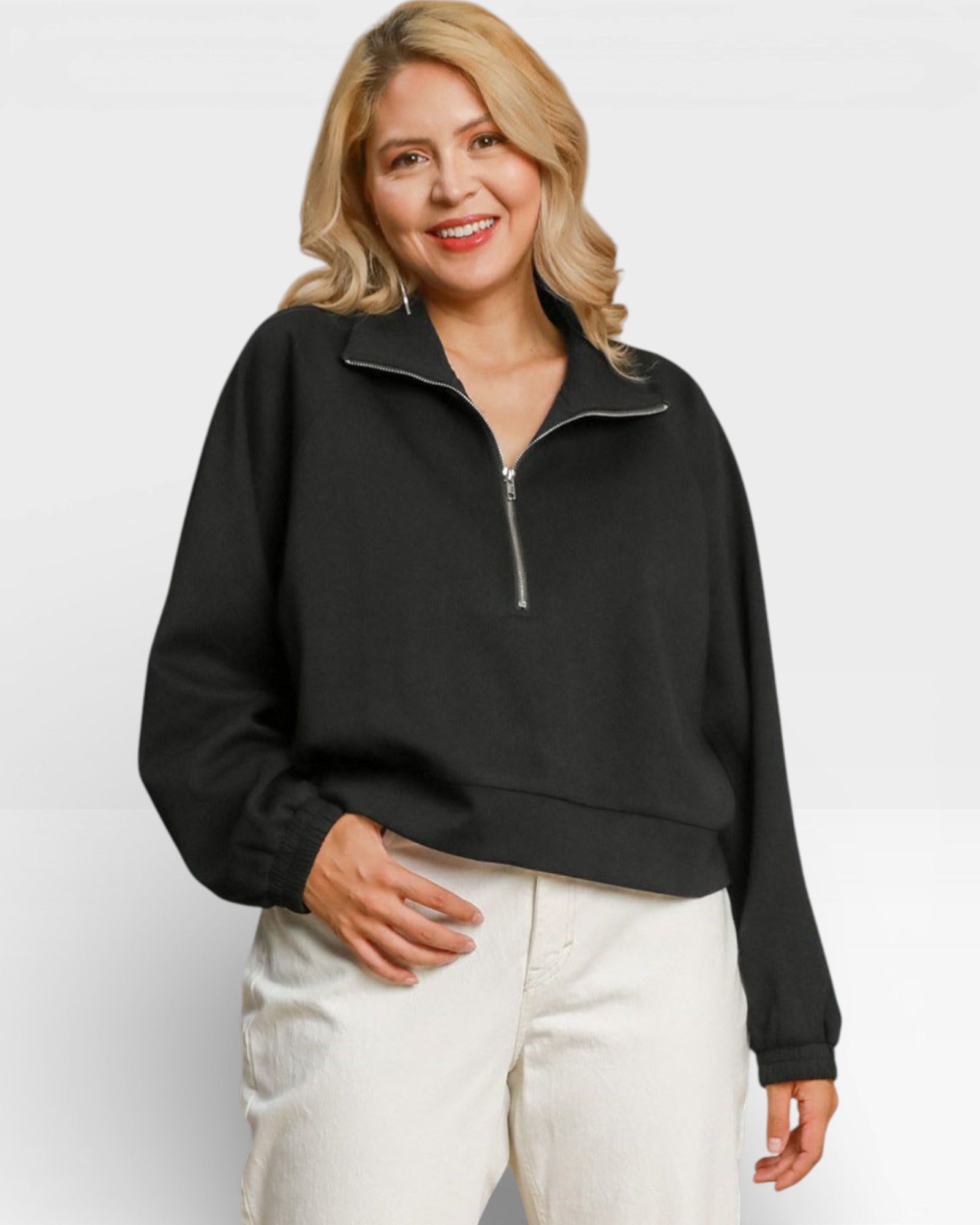 Woman wearing a black quarter-zip sweatshirt and white pants against a plain background