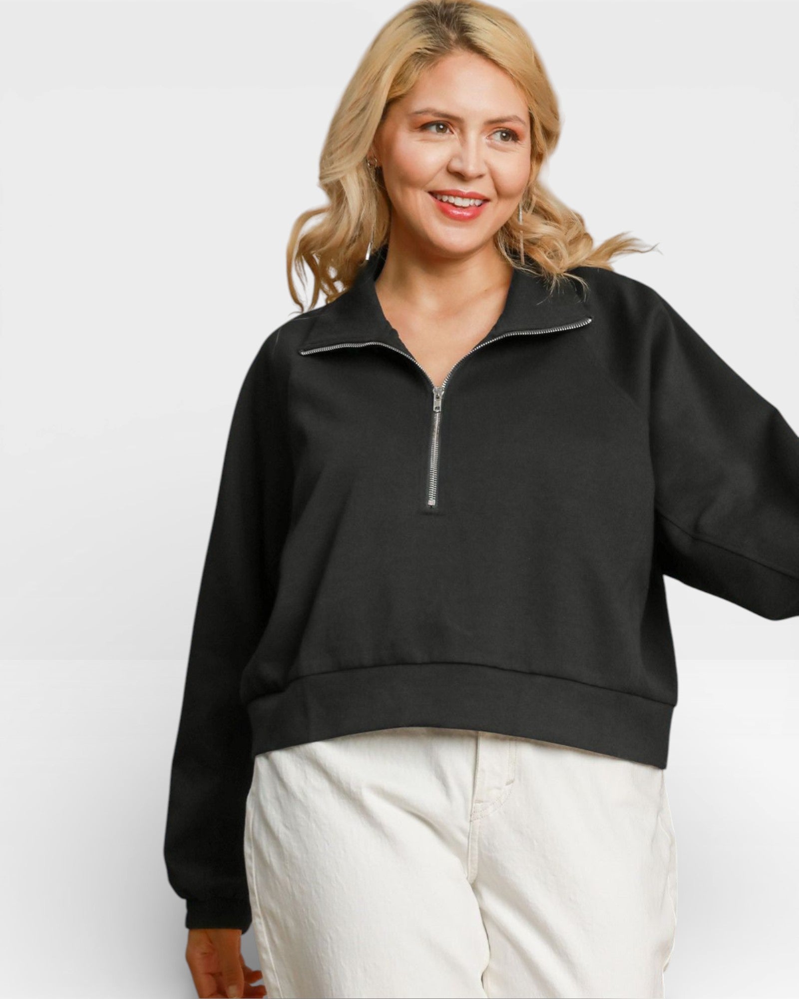 Woman wearing a black quarter-zip sweatshirt and white pants on a white background