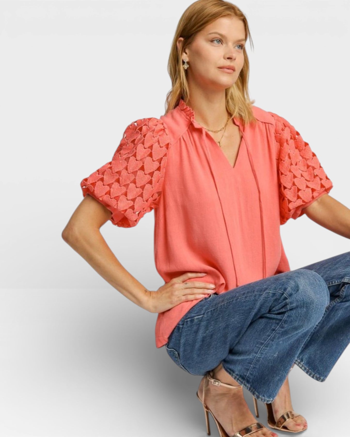 Woman wearing a coral blouse with ruffled sleeves and blue jeans on a light gray background