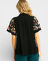 Black blouse with floral embroidery on sleeves worn by a person against a light gray background