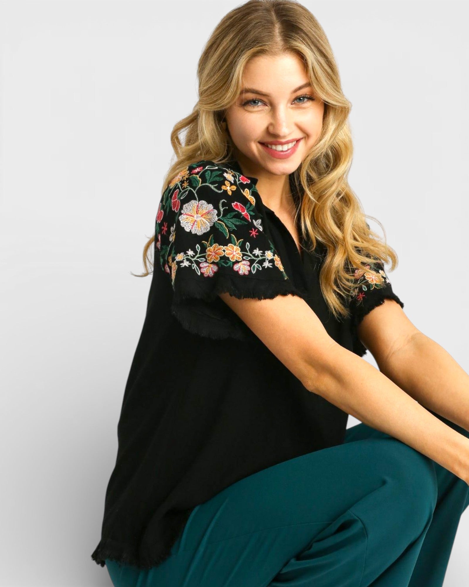Woman wearing a black top with floral embroidery and teal pants on a light gray background