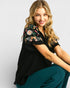 Woman wearing a black top with floral embroidery and teal pants on a light gray background