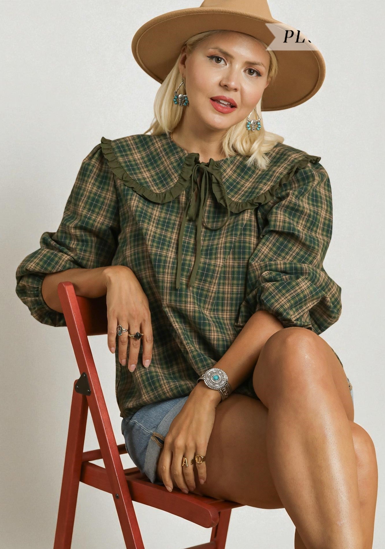 Woman wearing a green plaid shirt and beige hat sitting on a red chair.