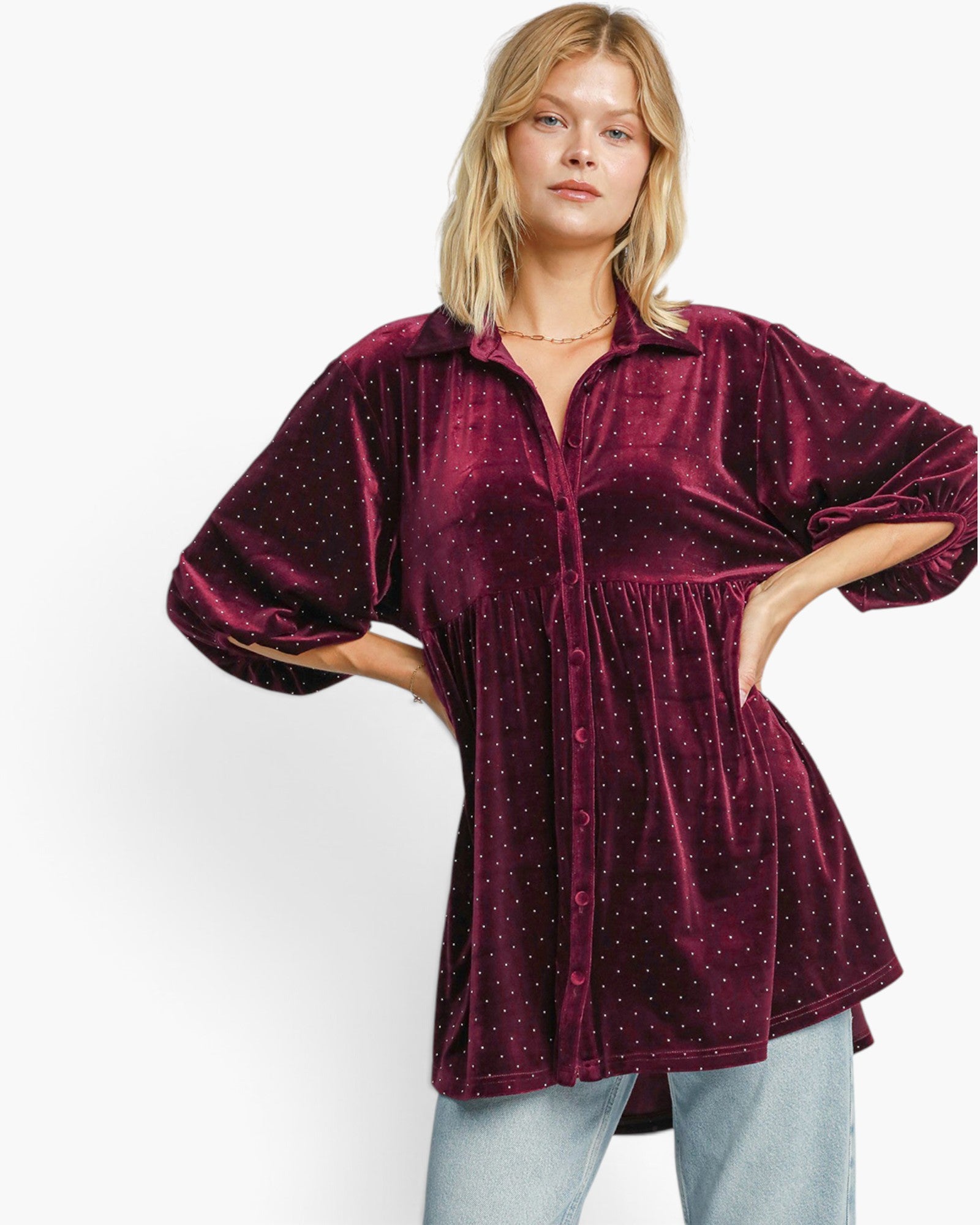 A woman modeling a deep burgundy velvet tunic top with a subtle gold polka dot pattern, paired with light blue jeans against a plain white studio background.