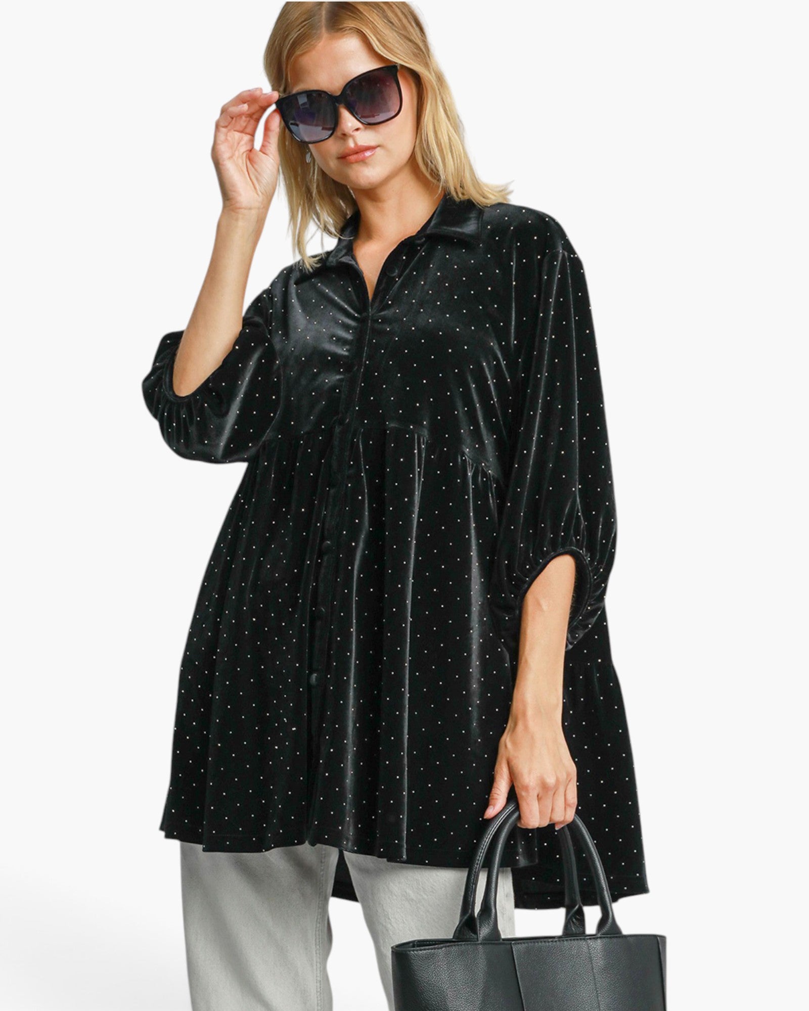 A woman modeling a black velvet long-sleeve tunic top with small white polka dots, styled with gray pants, black sunglasses, and a black tote bag against a plain white background.