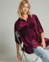 Woman wearing a burgundy velvet shirt sitting on a chair against a plain background