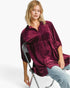 A woman wearing a burgundy velvet button-up shirt with tiny white polka dots, paired with light blue jeans, sitting on a black metal folding chair against a white studio background.